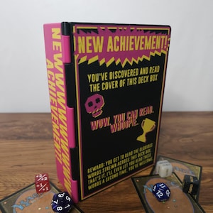 May include: A black and pink plastic deck box featuring yellow text that reads NEW ACHIEVEMENT! YOU'VE DISCOVERED AND READ THE COVER OF THIS DECK BOX. WOW, YOU CAN READ. WHOOPIE. REWARD: YOU GET TO READ THE GLORIOUS WORDS STREWN ACROSS THIS DECK BOX.