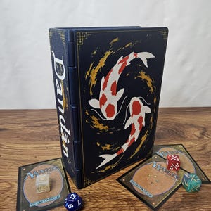 May include: A dark blue rectangular deck box featuring a white and red koi fish graphic and gold accents. The side of the box displays the word Dandan in white text. Two Magic the Gathering cards and three multi-colored dice are placed nearby.