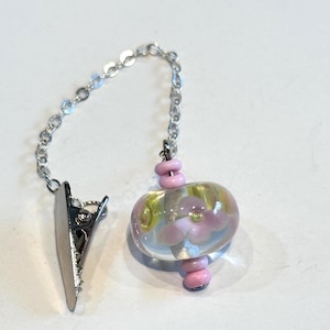May include: A silver-toned chain with a clip and a decorative bead. The bead is clear with pink and yellow floral designs and pink accent beads. The clip is attached to the chain.