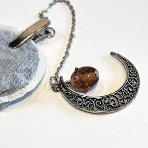 May include: A silver tea infuser with a crescent moon design and a decorative brown bead. The infuser has a chain and clip for attaching to a teacup. A tea bag is clipped to the infuser.