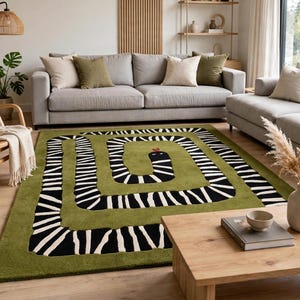Puede incluir: Una alfombra rectangular con un diseño de serpiente en espiral con rayas blancas y negras sobre un fondo verde oliva sólido. La serpiente tiene una pequeña lengua roja visible en la cabeza y la alfombra está sobre un suelo de madera clara.