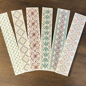 May include: Six rectangular bookmarks made of off-white cardstock, each featuring a unique geometric pattern embroidered with colorful thread. The designs include cross-stitch and linear motifs in shades of red, green, blue, and gold.