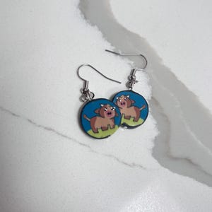 May include: A pair of round dangle earrings featuring a cartoon brown cow illustration on a blue and green background. The earrings are attached to silver-toned metal fishhook wires, designed for hanging from the earlobe with a simple hook mechanism.
