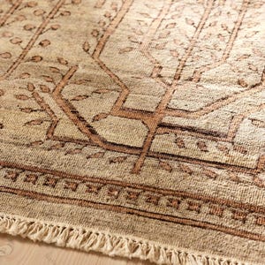 May include: A beige area rug featuring a repeating brown tree pattern with stylized branches and leaves. The rug has a decorative border with a geometric square motif and a fringe edge made of off-white knotted yarn along the bottom side.