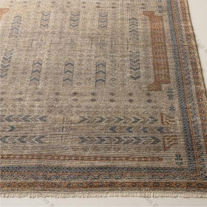 Peut inclure: Un tapis rectangulaire avec une base beige ornée de motifs répétitifs en chevrons bleus, de petites formes géométriques et de bordures marron. Le tapis présente un tissage texturé et une courte frange sur le bord inférieur du tissu.