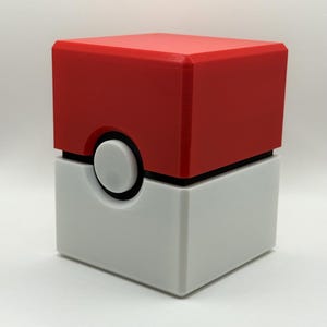 May include: A cubic container featuring a red top half and a white bottom half, separated by a thin black horizontal line. A circular white button is centered on the front, surrounded by a black ring, creating a distinct geometric design.