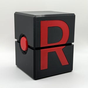 May include: A black cubic container featuring a large red letter R embossed on the front face. The cube is divided horizontally into two equal sections, with a small, circular red button positioned on the side seam of the structure.