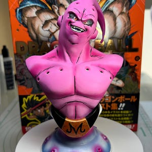 May include: A pink Majin Buu bust figurine featuring a black belt with a gold M emblem, set against a Dragon Ball orange box. The character has a smiling expression, dark eyes, and a curved head protrusion, mounted on a blue and white base.