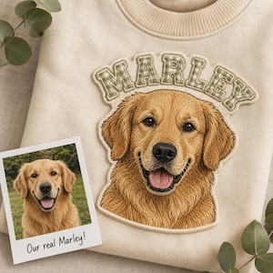 May include: A cream-colored sweatshirt featuring a large embroidered patch of a golden retriever's face with the name MARLEY in green and white gingham letters above it, accompanied by a small photograph labeled Our real Marley!