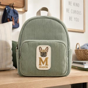 Peut inclure: Un sac à dos en velours côtelé vert sauge avec une poignée supérieure et une poche frontale zippée. La poche présente un écusson rectangulaire avec une illustration brodée de bouledogue français, un grand M doré et le nom MASON inscrit.