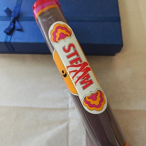Può includere: Un articolo in tessuto marrone scuro avvolto in una confezione di plastica trasparente. Un'etichetta bianca sulla parte anteriore presenta la parola STEAM in lettere rosse, circondata da un bordo decorativo smerlato arancione e rosa.