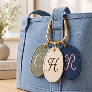 May include: A blue denim-textured bag featuring a gold-toned carabiner clip attached to three oval-shaped tags. The tags are olive green with a white letter C, cream with a brown letter H, and navy blue with a pink letter R embroidered on them.