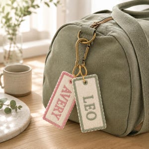 Può includere: Una borsa da viaggio in tela verde oliva con un moschettone in ottone attaccato alla cerniera. Due etichette rettangolari in feltro pendono dal moschettone, una con bordi rosa e il nome AVERY, l'altra con bordi verdi e il nome LEO.