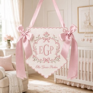 May include: A cream-colored fabric wall hanging with scalloped edges, featuring pink floral embroidery, a central monogram with the letters EGP, and the name Ella Grace Parker. The piece is suspended by a pink satin ribbon with bows on each side.