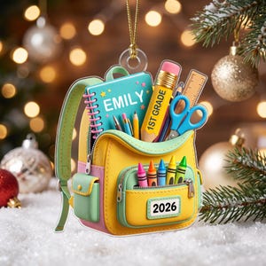 Puede incluir: Un adorno de mochila amarillo y verde claro con un cuaderno azul etiquetado EMILY, un lápiz amarillo etiquetado 1ST GRADE, una regla, tijeras y crayones de colores. El bolsillo frontal muestra el año 2026. Cuelga de un cordón dorado.