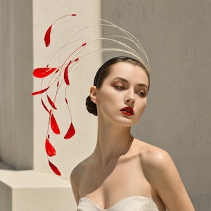 May include: A black headband fascinator featuring long, thin, arching stems that extend upward and outward. Each stem is tipped with a vibrant red feather, creating a dynamic, sweeping arrangement that curves gracefully above the head.