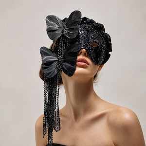 May include: A black masquerade mask featuring intricate lace detailing, a ruffled fabric trim, and two large black feather butterflies attached to the side. Long, delicate black lace ribbons hang down from the butterflies, adding texture and length.