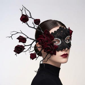 Könnte beinhalten: Eine schwarze Spitzenmaske mit silbernem Strassbesatz um die Augenöffnungen. Die Maske ist mit dunkelroten Rosen und schwarz bemalten Zweigen verziert, die seitlich herausragen und einen skulpturalen Kopfschmuck-Effekt erzeugen.