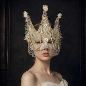 May include: A gold-toned lace masquerade mask shaped like a crown, featuring intricate embroidery, pearl-like embellishments at the points, and a fine mesh veil that drapes over the eyes and cheeks, creating a structured and ornate facial accessory.