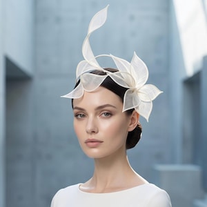 May include: A white fascinator headpiece featuring structured, mesh-like fabric petals arranged in a floral pattern. The design includes a tall, curved, leaf-like element extending upwards and several layered, pointed petals attached to a thin headband.