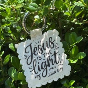 May include: A square off-white fabric keychain with scalloped edges featuring the black printed text Jesus IS THE light JOHN 8:12. The keychain is attached to a silver metal ring and displays decorative black flourishes around the lettering.