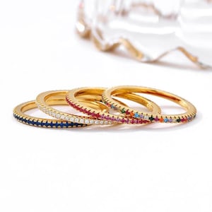 Puede incluir: Cuatro anillos finos de oro dispuestos en fila, cada uno con un canal de pequeñas piedras preciosas. Los anillos muestran piedras azules, transparentes, rojas y multicolores, engastadas en una banda metálica con borde estriado.