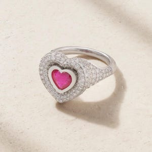 Könnte beinhalten: Ein silberfarbener Ring mit einem markanten herzförmigen Mittelstück. Die Mitte enthält einen leuchtend pinken herzförmigen Stein, der von einem weißen Rand umgeben ist, welcher wiederum von mehreren Reihen kleiner, klarer Glitzersteine eingefasst ist.