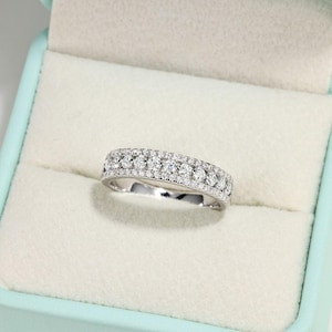 May include: A silver-coloured band ring featuring a central row of round, clear stones flanked by two thinner rows of smaller, pavé-set clear stones. The ring is crafted with a polished metal finish and displays a multi-row design.