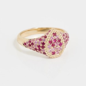 Könnte beinhalten: Ein goldener Ring mit einem birnenförmigen Mittelstück, das von einem gedrehten Metallrand umgeben ist. Die Oberfläche des Rings und die zentrale Birnenform sind dicht mit zahlreichen kleinen, runden, kräftig rosa und hellrosa Edelsteinen besetzt.