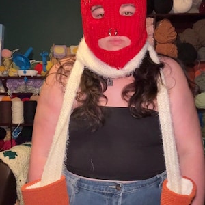 May include: A red crocheted balaclava shaped like a strawberry with a green stem on top. The headpiece features a white fuzzy scarf integrated into the design, extending downwards with two orange textured mittens attached to the ends of the scarf.