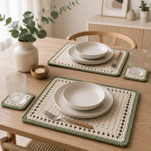 May include: A set of rectangular crochet placemats and square coasters featuring a cream-coloured centre with a decorative open-stitch border and a solid sage green edge. The placemats are topped with white ceramic plates and silver forks.