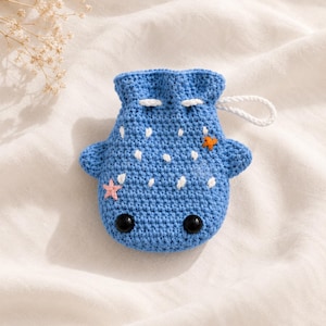 Può includere: Un sacchetto in crochet blu con coulisse a forma di squalo balena, caratterizzato da punti bianchi ricamati, due occhi in plastica nera, una piccola applicazione a forma di stella marina rosa, una arancione e una chiusura con cordino bianco.