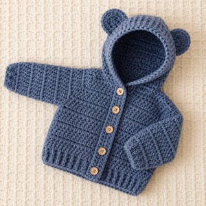 May include: A blue long-sleeve crochet cardigan featuring a hood with two rounded bear-like ears. The garment has a front button closure with five light-coloured wooden buttons and a ribbed hem at the bottom and on the sleeve cuffs.