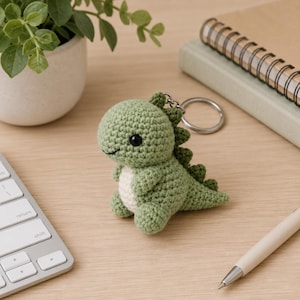 May include: A small, light green crocheted dinosaur keychain with a cream-colored belly, black bead eyes, and a row of dark green spikes along its back. The plush figure is attached to a circular silver metal keyring for easy attachment to bags.