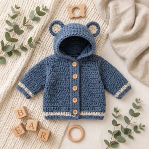 Peut inclure: Un gilet à capuche en crochet bleu avec des oreilles d'ours sur la capuche, cinq boutons en bois brun clair sur le devant, et des rayures crème sur les poignets et l'ourlet inférieur, réalisé avec un motif de maille texturé.