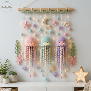 May include: A wall hanging featuring three crocheted jellyfish in pink, mint green, and lavender, suspended from a wooden rod with a green wavy trim. The display includes crocheted stars, shells, small spheres, a crescent moon, and string lights.