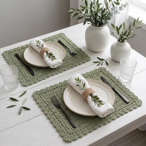 May include: Two rectangular sage green crocheted placemats with scalloped edges, each featuring a white ceramic plate, a white cloth napkin held by a woven ring, a black fork, a black knife, and a clear ribbed glass on a white wooden table.