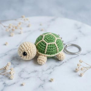 Pode incluir: Um porta-chaves de tartaruga em croché com cabeça e membros de cor creme e uma carapaça verde com costuras em cor creme. A tartaruga tem pequenos olhos de missangas pretas e está presa a um anel metálico de tom prateado.