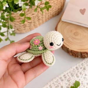 May include: A small, handmade crochet turtle toy featuring a cream-coloured head and limbs with a sage green shell decorated with small pink and yellow floral appliques. The turtle has black bead eyes and stitched pink cheeks with a simple black smile.