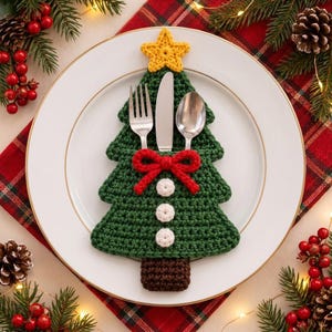 May include: A dark green crochet Christmas tree cutlery holder featuring a yellow star at the top, a red bow, and three white circular accents. The tree-shaped pocket contains a silver fork, a silver knife, and a silver spoon.