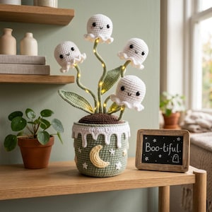 May include: A crocheted plant featuring four white ghost-shaped flowers with black eyes and smiles, illuminated by small lights on green stems. The plant sits in a sage green crocheted pot decorated with a yellow crescent moon and white stars.