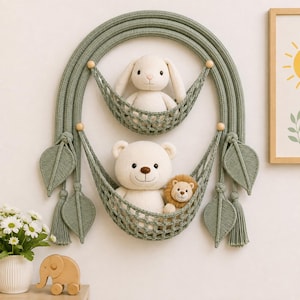 May include: A sage green macrame wall hanging featuring a large arched frame with two integrated net hammocks. The top hammock holds a cream-colored plush rabbit, while the bottom hammock contains a cream plush bear and a small brown plush lion.