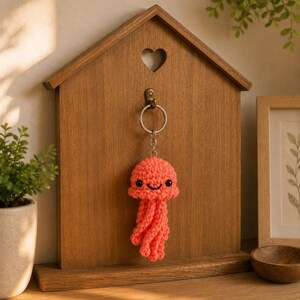 May include: A coral-colored crochet jellyfish keychain featuring a rounded head, black plastic safety eyes, a stitched black smile, and multiple dangling tentacles. The jellyfish is attached to a silver-toned metal split ring keychain.