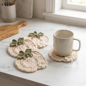 May include: Three round, cream-colored crochet coasters shaped like pumpkins, each featuring a textured green stem and a small white daisy with a yellow center. A speckled, off-white ceramic mug sits on a fourth matching coaster.