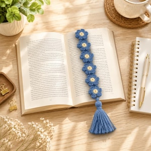 May include: A handmade crochet bookmark featuring a vertical row of six blue flowers with yellow centers, finished with a matching blue tassel at the bottom. The bookmark is placed inside an open book with printed text on the pages.