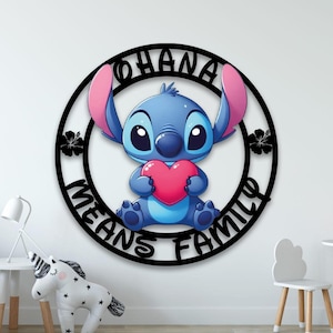 Può includere: Un'insegna da parete circolare nera con un personaggio alieno blu che tiene in mano un cuore rosa. Il testo "OHANA MEANS FAMILY" è integrato nella cornice circolare, accompagnato da due sagome di fiori di ibisco neri ai lati.