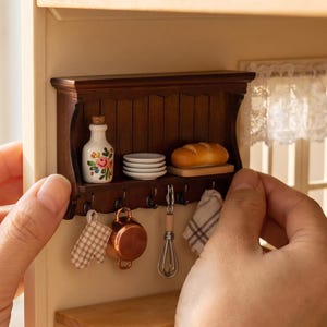 Puede incluir: Una estantería de pared en miniatura de madera marrón oscuro para cocina de casa de muñecas, con una repisa superior con una botella floral, una pila de platos blancos y un pan, además de cinco ganchos metálicos con un guante de cocina y un batidor.