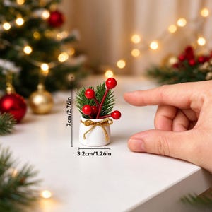 Könnte beinhalten: Ein Miniatur-Topf aus weißer Keramik mit grünen Tannennadeln und roten Beeren, verziert mit einer goldenen Schnurschleife. Der Artikel misst 7 cm in der Höhe und 3,2 cm in der Breite, wie durch die Maßangaben deutlich wird.