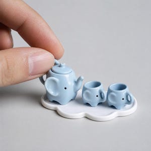 May include: A miniature light blue ceramic tea set featuring an elephant-shaped teapot and two matching elephant-shaped cups, all arranged on a small white cloud-shaped tray. Each piece has small black painted eyes and elephant ears.