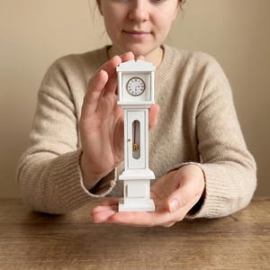 May include: A miniature white grandfather clock featuring a classic rectangular frame, a circular clock face with Roman numerals, and a central glass panel revealing a thin red pendulum with a gold-coloured weight at the bottom of the clock body.